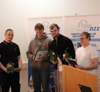 Vier Auszubildenden stehen vor den RollUps des Ausbildungszentrum Zwickau, in den Händen halten Sie ihre Abschlusszeugnisse und Blumen. 
