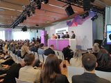 Blick vom gut besuchten Besuchersaal auf das Podium, auf dem fünf Gesprächspartner*innen diskutieren.