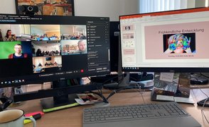 Fotografiert wurde ein Schreibtisch mit zwei Computerbildschirmen. Auf dem linken Bildschirm ist die Videokonferenz zu sehen, auf dem rechten Bildschirm läuft die Powerpoint-Präsentation zum Fachtag.