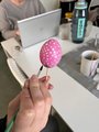 Zu sehen ist eine Hand, die ein auf ein Holzstäbchen gestecktes rosa Osterei mit weißen Punkten verziert.