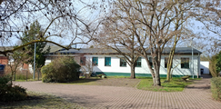 Foto: Einstöckiges Gebäude mit weiß-grüner Fassade, gepflastertem Vorplatz und kahlen Bäumen in einer ruhigen Außenanlage.