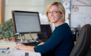 Eine Frau sitzt im Büro am Computer und lächelt in die Kamera.
