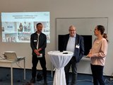 Foto: Drei Personen stehen hinter einem Stehtisch. An der Wand hinter ihnen ist eine Projektion zur Veranstaltung zu sehen. 