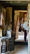 Eine junge Frau mit erhobenem Daumen sieht durch die Tür des Bauernhauses, im Vordergrund alte Bauernhof-Gerätschaften sowie trockene Räucherware.