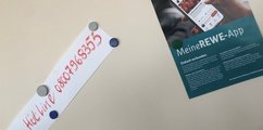 Foto einer Pinnwand, an der ein Poster mit der Aufschrift Meine REWE-App sowie ein handgeschriebener Zettel mit Magneten befestigt sind.