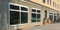 Foto: Gebäudefassade mit großen Schaufenstern, Pflanzkübeln vor dem Eingang und mehreren abgestellten Fahrrädern am Gehweg.