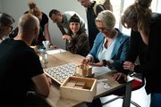 Viele Personen sitzen und stehen um einen Tisch mit einem Holzbrett und probieren sich im kognitiven Training