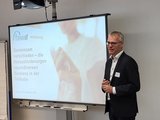 Foto: Reha Fachbereichsleiter, Herr Hagen, begrüßt lächelnd neben einem Smartboard stehend die Teilnehmenden
