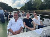 Foto von der Schifffahrt. Mehrere Teilnehmende sitzend und stehend auf dem Oberdeck. 
