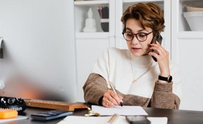 Eine Frau sitzt am Schreibtisch und macht sich beim Telefonieren Notizen.