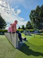 Sommerfest 2026 2 Mitarbeiter sitzen auf einer Betonmauer und schauen zu. Im Hintergrund sind einige Spielfelder, blauer Himmel, Wolken und Bäume erkennbar.