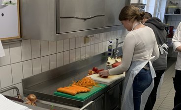 Ein Mädchen und ein Junge im Alter von 14 bis 15 Jahren stehen in einer Küche an einer Arbeitsplatte und schneiden Gemüse.