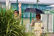 Zwei Mitarbeiterin lachen in die Kamera, die linke ist nur halb durch einen Busch zu sehen. Die rechte Frau hält einen Schirm als Sonnenschutz.