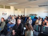 Plenum in Mainz Foto: Menschen in einem Seminarraum und folgen einer Veranstaltung, hinten steht ein Tisch mit Kaffee.