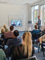 Foto: Menschen sitzen in einem Veranstaltungsraum und schauen auf einen Bildschirm in dem hybrider Unterricht gezeigt wird.