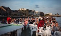 Foto vom Oberdeck des Schiffes auf die Teilnehmenden, dem Fluss und den umliegenden Gebäuden.