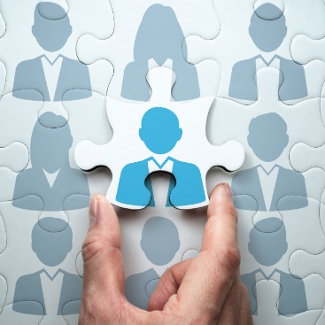 Eine Hand legt ein Puzzleteil mit einer Person zwischen weiteren, ähnlichen Teilen ab: Symbolbild Vermittlung & Beratung