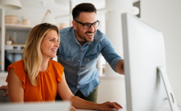 Qualifizierungssituation: Mann steht neben sitzender Frau, beide schauen auf den Monitor; der Mann zeigt auf den Bildschirm.