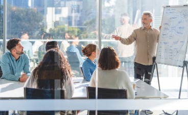 Beispielfoto: fünf Menschen sitzen vor großer Fensterfront um einen Tisch, den Blick auf den Redner am Whiteboard gerichtet. 