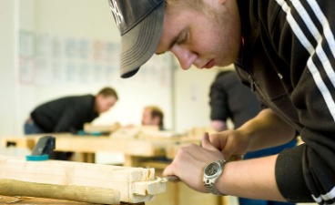 Foto: Konzentriert arbeitet ein Mensch an einem Holzstück. Im Hintergrund verschwommen zwei weitere Personen bei der Arbeit. 