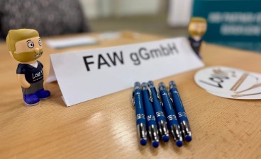 Foto: Werbemittel, wie Stifte, Aufkleber sowie ein faw gGmbH-Namensaufsteller, eingerahmt von zwei kleinen Lou!s-Spielfiguren.