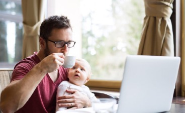 Ein Mann sitzt am Tisch vor einem Laptop mit einem Baby auf dem Schoss und trinkt Kaffee