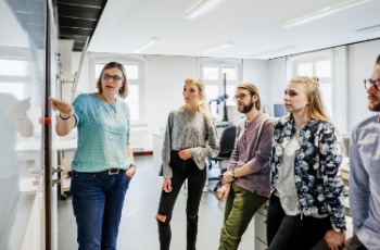 Beispielbild Coachingsituation: Vier Personen stehen um eine Person, die am Whiteboard etwas erklärt. 