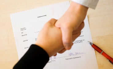 Auf einem Tisch liegt ein unterschriebener Arbeitsvertrag. Über den Tisch hinweg geben sich Arbeitgeber und Arbeitnehmer einen Handschlag.