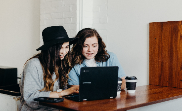 Zwei junge Frauen sitzen vor einem Laptop und beraten über das, was sie sehen.