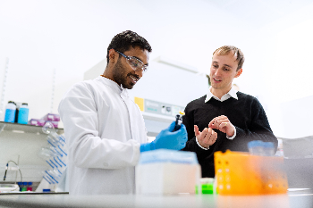 Zwei Männer stehen in einem Labor, einer trägt weiße Schutzkleidung und Handschuhe. Er hält einen kleinen Behälter für Flüssigkeiten in der Hand, der andere erklärt dem ersten etwas.