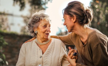 Eine Frau sitzt neben einer Rentnerin, hält ihre Hand und unterhält sich mit ihr.