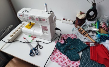 Foto: Nähtisch, auf dem links zwei Stoffscheren vor einer Nähmaschine liegen, daneben verschiedene Stoffstücke.