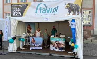 Foto: Pavillon in dem drei Menschen, Stehtische und verschiedene Roll-Ups zu sehen sind. Darüber ein großes faw-Banner.