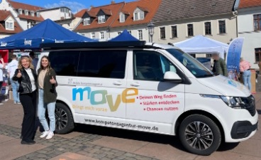 Ein weißer Bus mit Aufdrucken der Kompetenzagentur MOVE steht auf einem Marktplatz. Links daneben stehen zwei Kolleginnen des Projektteams.