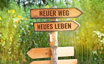 Auf einer Wiese steht ein Wegweiser. Zwei Richtungspfeile zeigen in die gleiche Richtung. Der eine Pfeil ist beschriftet mit „Neuer Weg", der andere mit „Neues Leben".