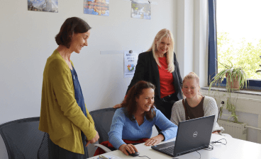 Foto: Vier Frauen am Laptop, zwei sitzend, zwei stehend dahinter. 