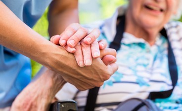 Beispielfoto: Eine Person hält mit beiden Händen die rechte Hand einer lächelnden Seniorin, welche in einem Rollstuhl sitzt.