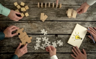 Loesungsdenker, gemeinsames Brainstorming Auf einem Tisch liegen Puzzelteile und Holzklötze sowie ein Bild von einer Glühbirne. Dazu sind mehrere Hände zusehen. Es werden Ideen entwickelt.