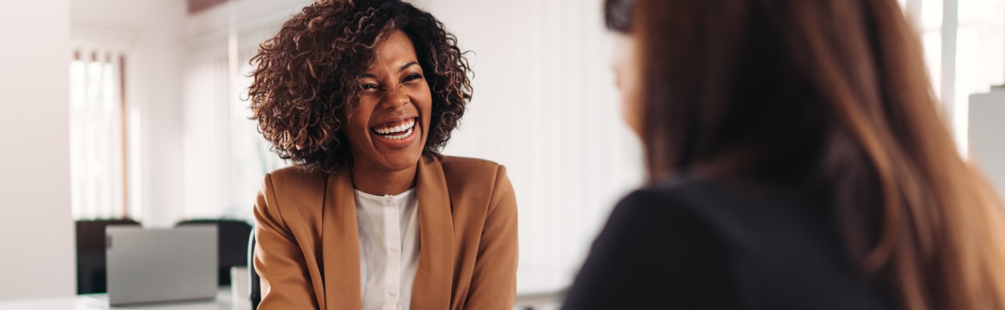 Symbolbild: Zwei Frauen sitzen sich lachend an einem Schreibtisch gegenüber