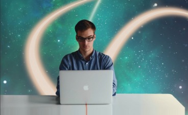 Mann mit Brille sitzt an weißem Schreibtisch vor seinem weißen Laptop. Hinter ihm an der Wand ist das Weltall in blautürkis abgebildet.