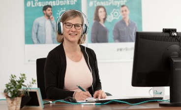 Beispielfoto: Frau mit Kopfhörer sitzt vor einem PC Bildschirm und lächelt in die Kamera