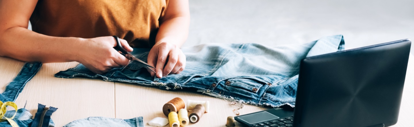 Beispielbild: Person schneidet an einer Jeans. Auf dem Tisch außerdem Fadenrollen, Maßband u. weitere Nähutensilien sowie Laptop.
