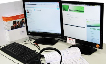 Das Bild zeigt einen Arbeitsplatz für ein digitales Lernangebot mit 2 Monitoren, Headset, Tastatur und Arbeitsmaterialien