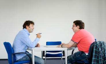 Dargestellt ist ein Foto, auf dem man zwei Herren sieht, die an einem Tisch sitzen und miteinander sprechen. Einer der beiden macht sich Notizen.