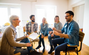 Beispielfoto Gesprächssituation: Fünf Personen sitzen in einem hellen Raum auf Stühlen im Kreis und sprechen miteinander.