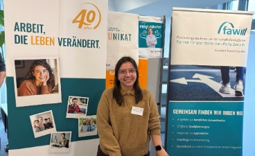 Melanie Detzel steht lächelnd vor dem Infostand auf der Bildungsmesse in Landau im Jahr 2026. Links befindet sich ein Roll‑Up mit der Aufschrift ‚Arbeit, die Leben verändert. 40 Jahre‘ sowie mehreren Fotos von Menschen. Rechts steht ein weiteres Roll‑Up der FAW Kaiserslautern mit Informationen zu Bildungs- und Unterstützungsangeboten. Im Hintergrund sind weitere Messebanner und ein heller Innenraum zu sehen.