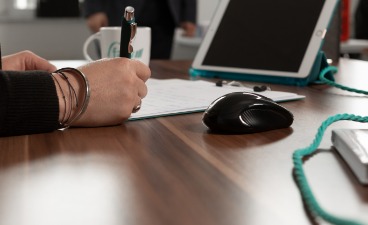 Beispielbild Arbeitsplatz: Schreibtisch mit Bildschirm und Computermaus, an dem eine Person, von der nur die Hand zu sehen ist, sitzt und etwas schreibt.