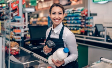 Beispielfoto: Person in schwarzer Schürze scannt an der Kasse lächelnd eine Produkt ein. nd Regale mit Feinkostprodukten. 