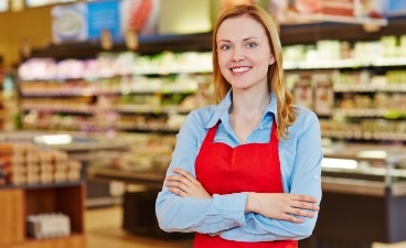 Beispielfoto: Junge Verkäuferin mit roter Schürze steht mit verschränkten Armen lächelnd vor Regalen im Supermarkt.