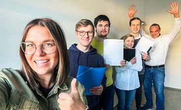 Foto: Sechs Personen lachen in die Kamera. Eine mit Daumen nach oben, ein anderer mit beiden Händen in der Höhe.